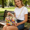 A young woman sits on a wooden bench holding a multi-color Oxford cloth picnic lunch bag.