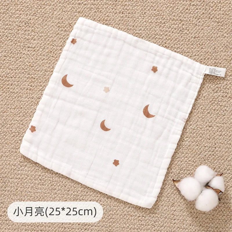 A white muslin baby burp cloth towel with small rose gold moon patterns is shown on a beige textured surface.