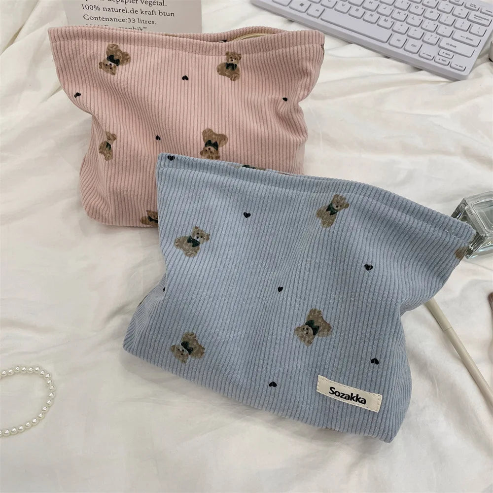 A light pink corduroy clutch bag with small brown bear embroidery resting on a white surface.