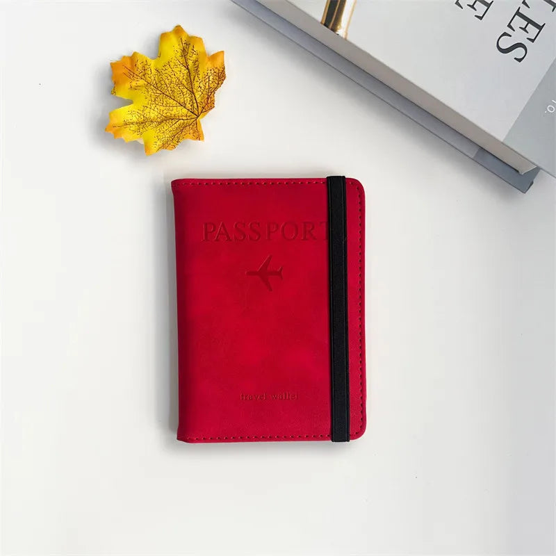 A red passport wallet with a black trim rests on a white surface next to a yellow autumn leaf and a book.