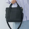 A woman holds a black canvas crossbody bag with an adjustable strap and multiple pockets against a white shirt and blue jeans