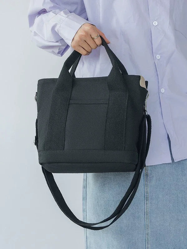 A woman holds a black canvas crossbody bag with an adjustable strap and multiple pockets against a white shirt and blue jeans