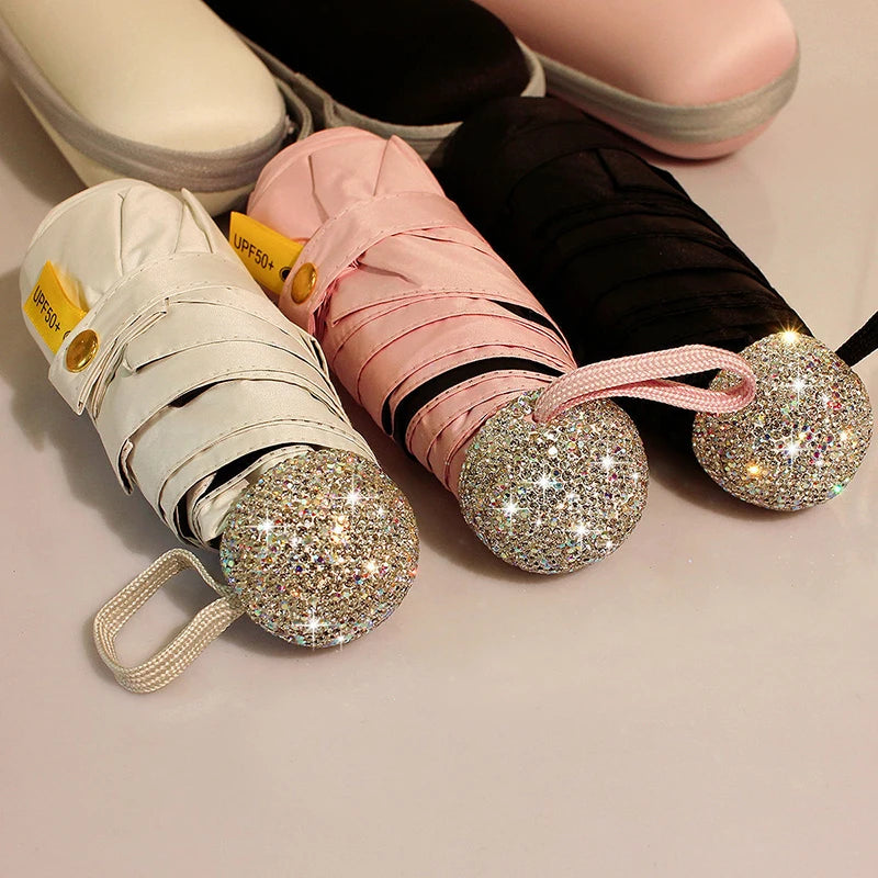A collection of compact rhinestone umbrellas in white and pink, some with black fabric, displayed on a light surface.