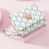 A light gray letter zipper wristlet wallet with a gold monogram detail resting on a pink surface.