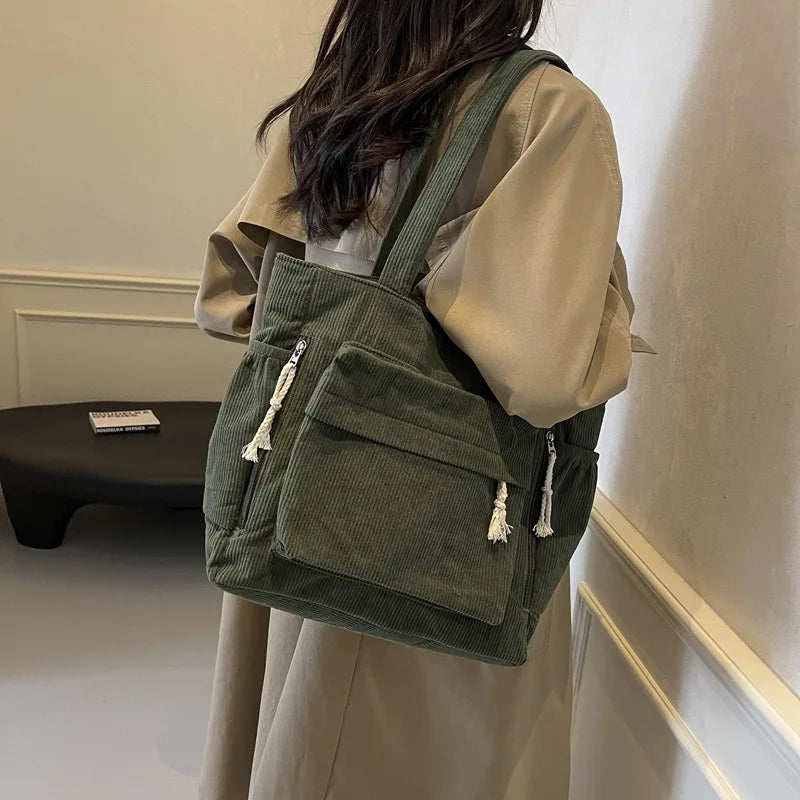 A person is holding a olive green corduroy shoulder bag with zipper details against a light background.