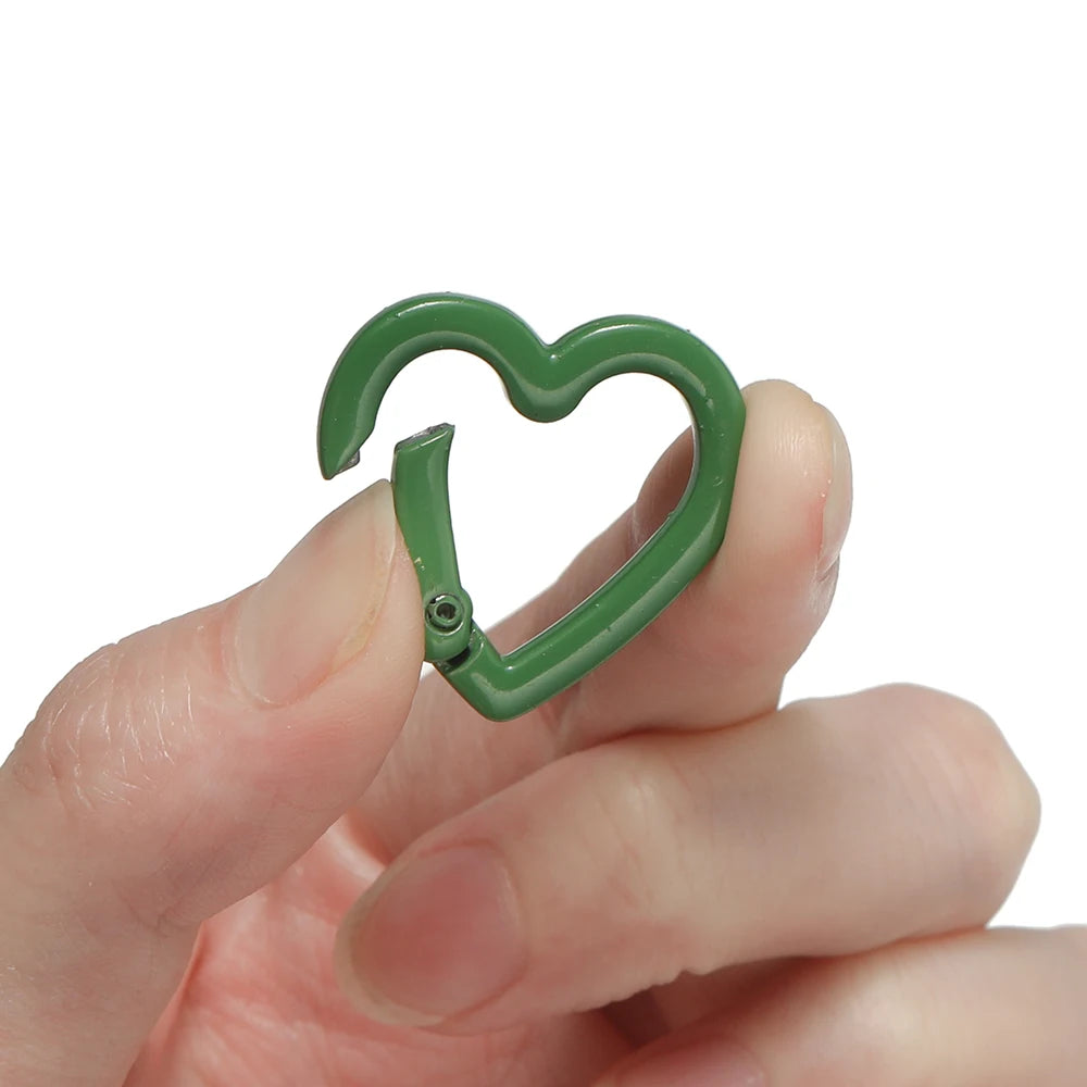 A hand holds a green heart-shaped metal spring clasp for DIY jewelry or keychains.