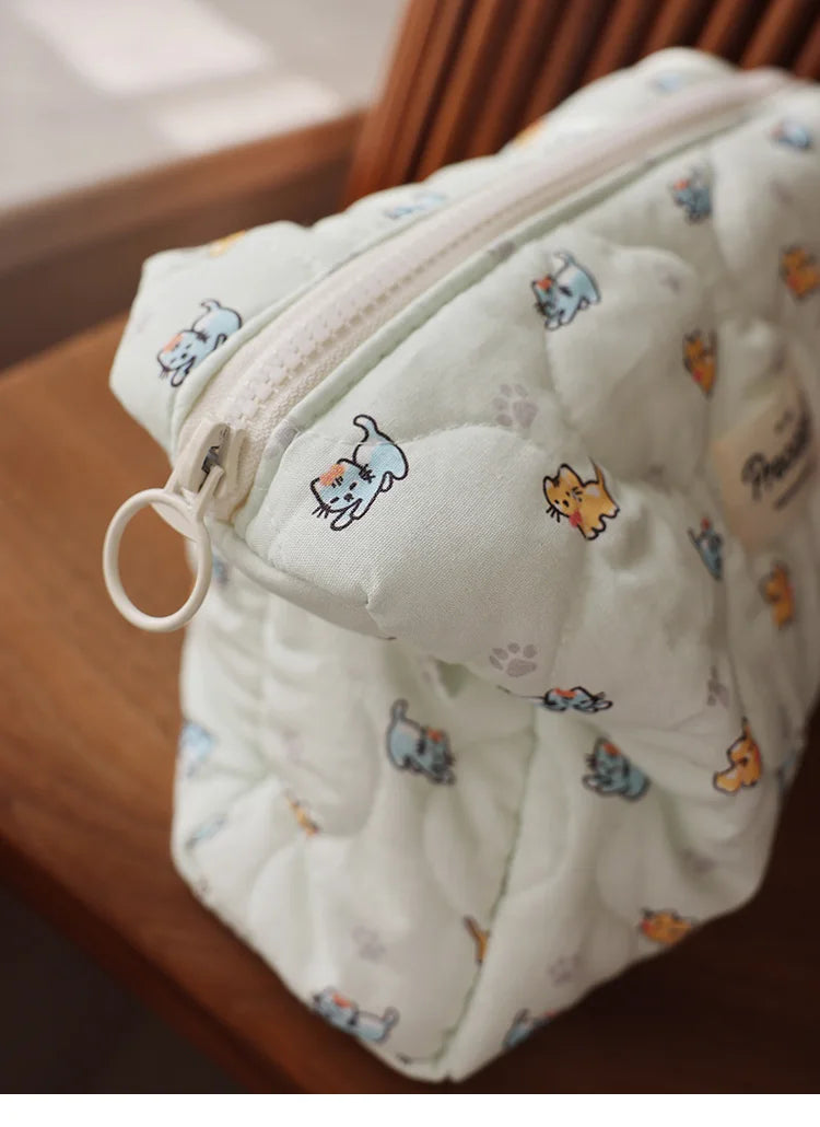 A quilted white cosmetic storage pouch with a zipper and small printed cats resting on a wooden surface.