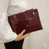 A close-up shot of a dark burgundy alligator print clutch bag being held by a person wearing a white shirt.