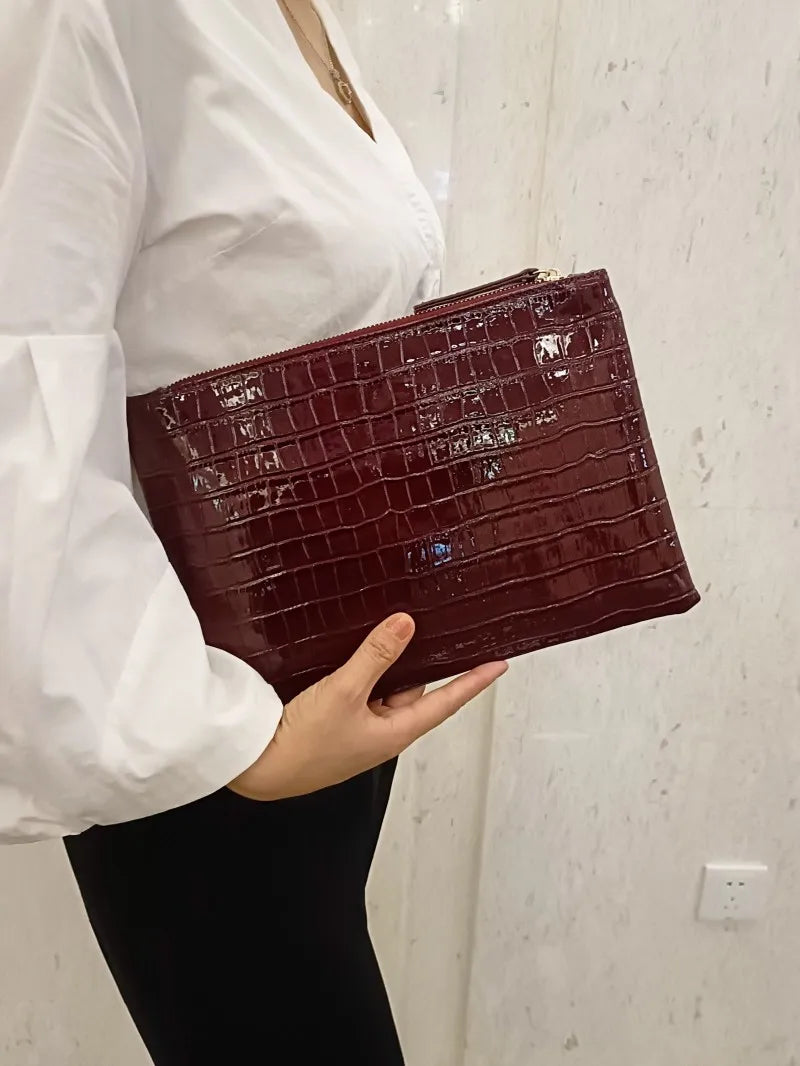 A close-up shot of a dark burgundy alligator print clutch bag being held by a person wearing a white shirt.