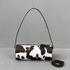 A brown and white cow print cylinder shoulder bag with a black strap is shown against a light gray background.