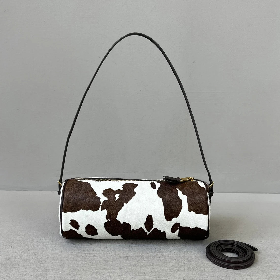 A brown and white cow print cylinder shoulder bag with a black strap is shown against a light gray background.
