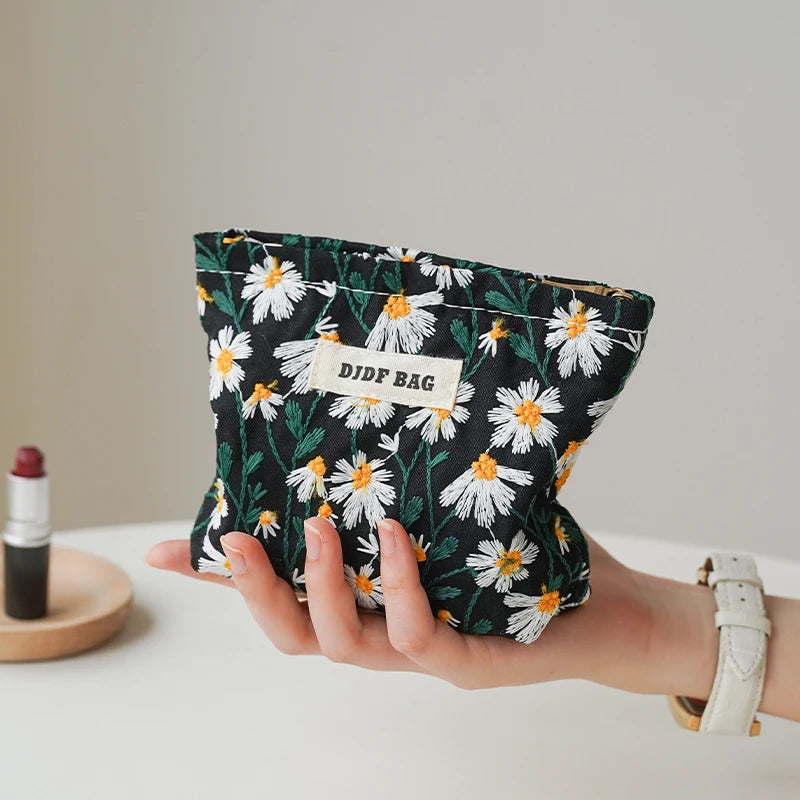 A hand holds a floral canvas makeup bag with white daisies and green leaves against a light background.