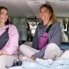 Two women sitting inside a van looking at the camera with bags and blankets nearby