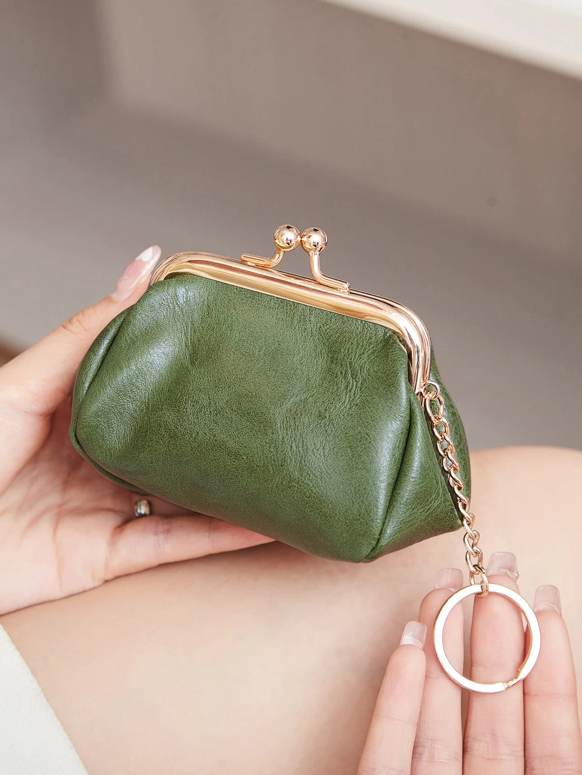 A hand holds a olive green leather coin purse with a gold chain keychain against a neutral background.