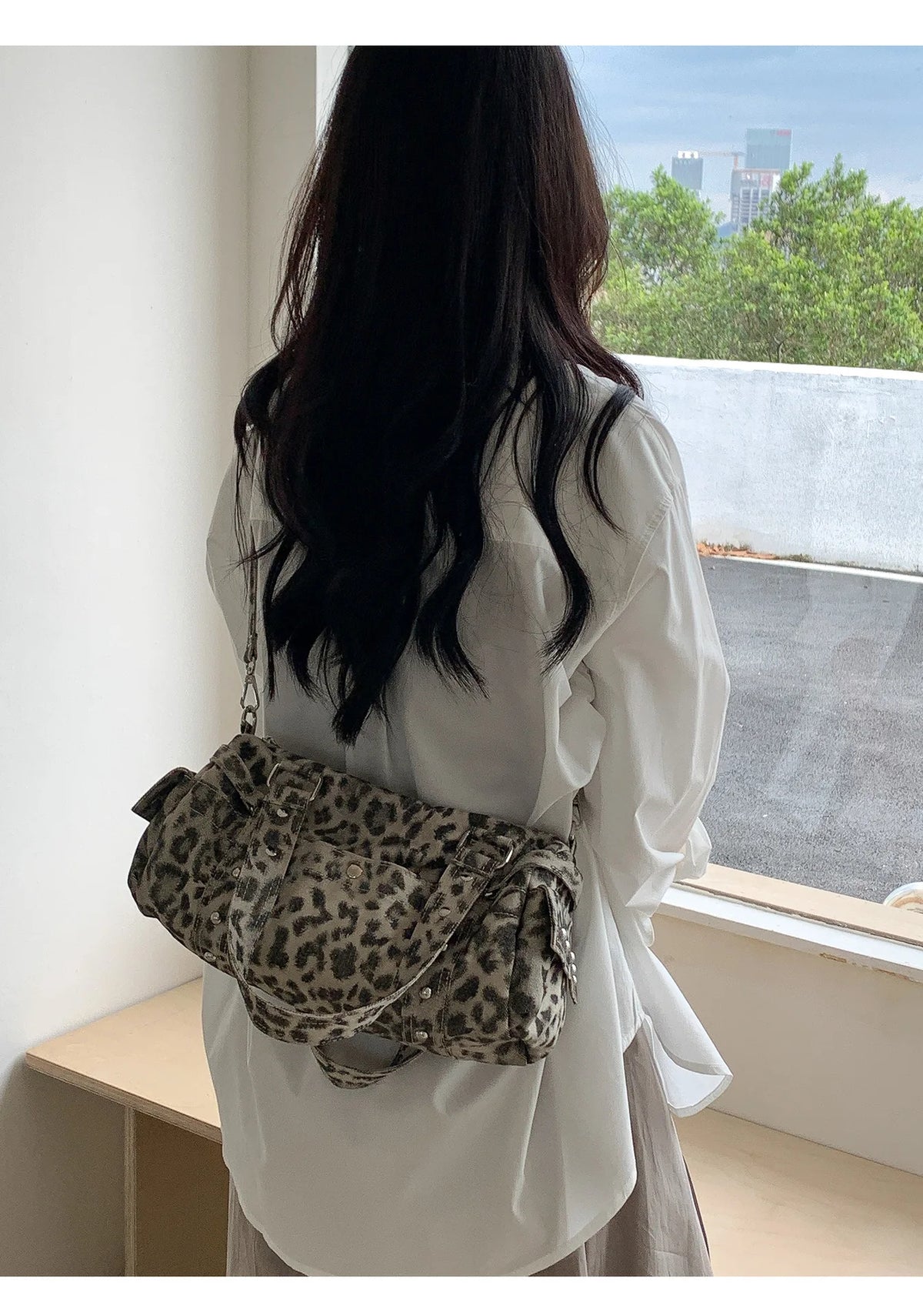 A woman stands by a window holding a leopard print riveted shoulder bag.