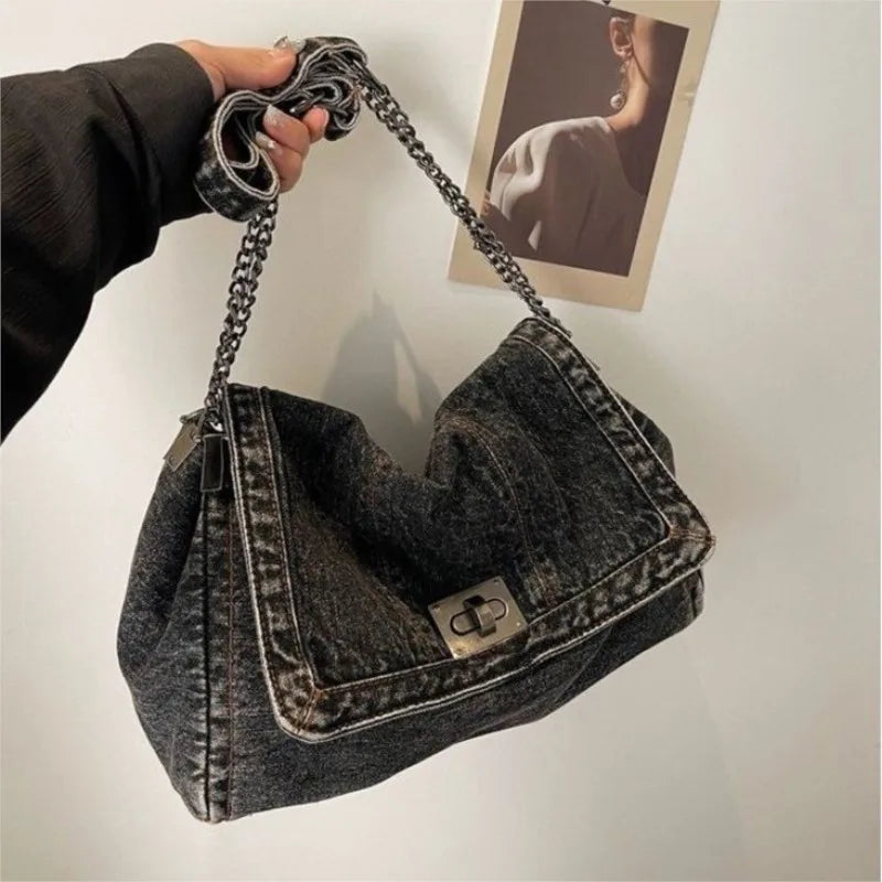 A dark denim tote bag with a chain strap is being held against a white wall.