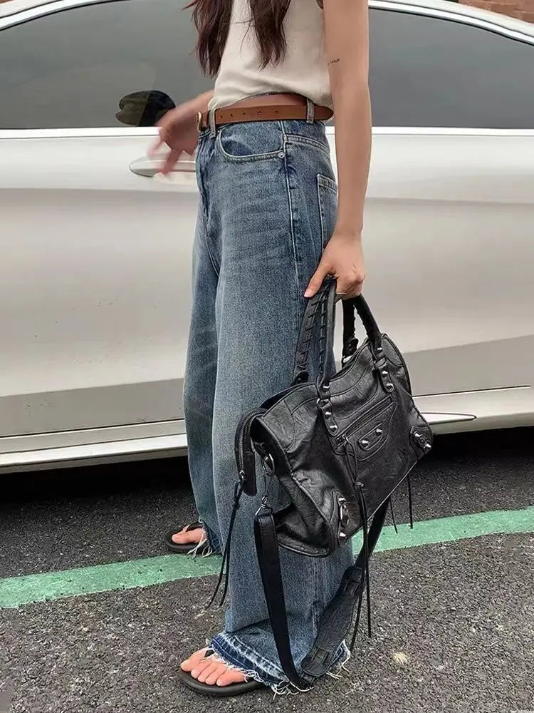 A woman wearing a white top and jeans stands next to a silver car, holding a black saddle-shaped PU leather city tote bag.