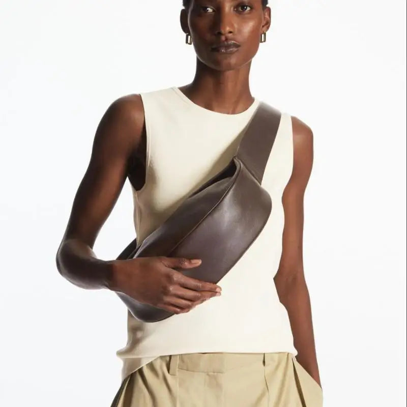 Una modelo vistiendo una camiseta sin mangas color crema y pantalones caqui, sosteniendo un bolso cruz de cuero marrón oscuro