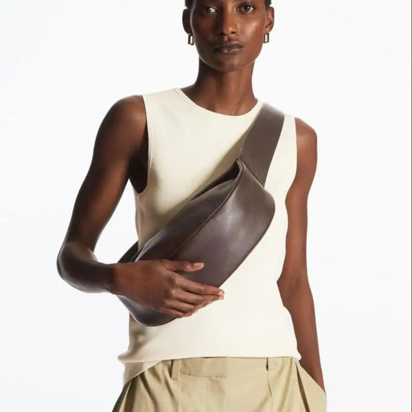 A model wearing a cream tank top and tan pants holding a dark brown leather crossbody bag.