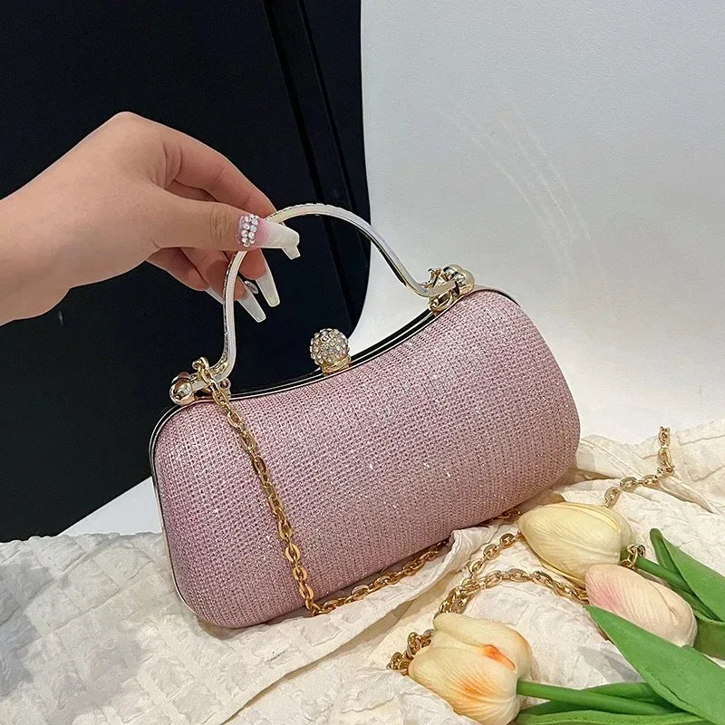 A hand holds a light pink glitter crossbody evening clutch bag with gold chain details on a white fabric surface.