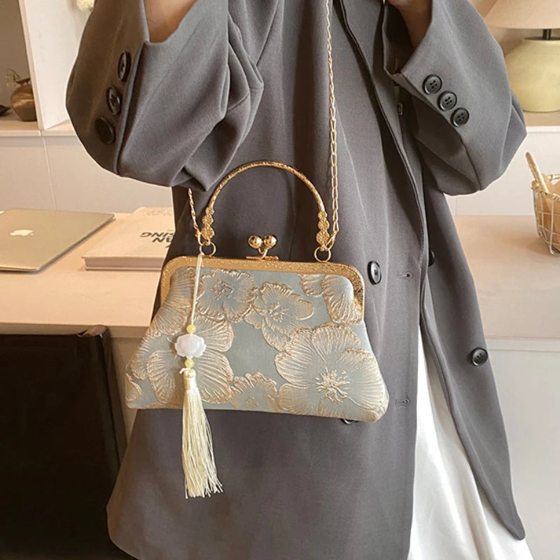 A light blue embroidered silk evening clutch with a gold chain strap is displayed against a gray coat.