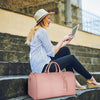 A woman wearing a straw hat is sitting on stone steps, looking at a tablet while resting a pink foldable multi-function garme