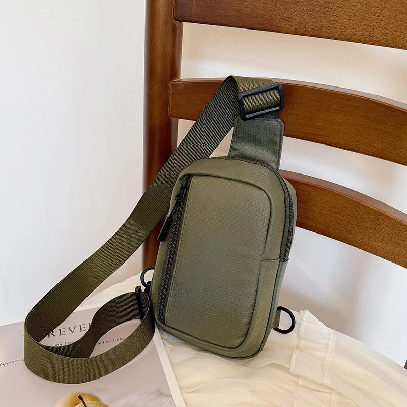 Olive green chest bag with a dark strap resting on a wooden chair.