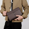 A person holds a dark brown men's PU leather clutch bag against a beige jacket.