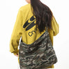 A person wearing a yellow sweatshirt and black beanie is holding a canvas camouflage shoulder bag.