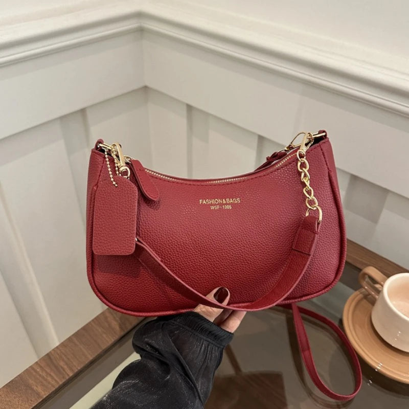 A maroon leather shoulder bag with a gold chain pendant resting on a wooden surface.