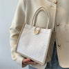 A beige blazer sleeve holds a natural linen tote bag with white floral embroidery and a wooden closure.