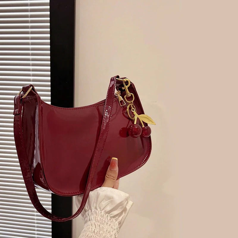 A maroon PU leather shoulder bag with a gold chain and cherry pendant is being held against a light wall.