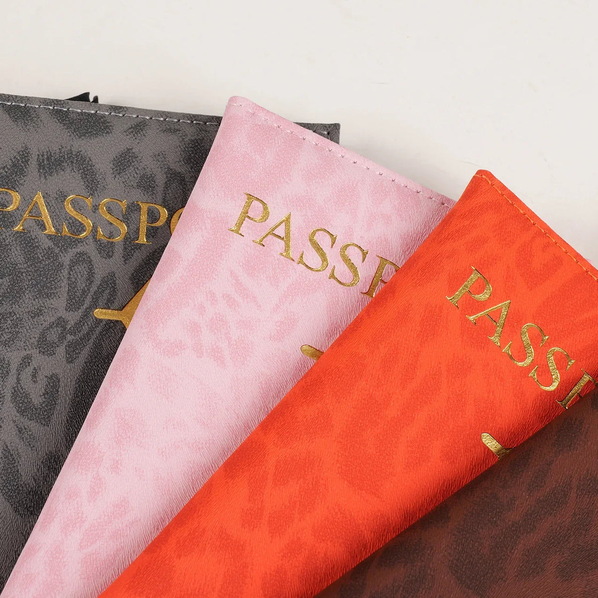 Three passport holders in shades of gray, pink, and orange with gold lettering.