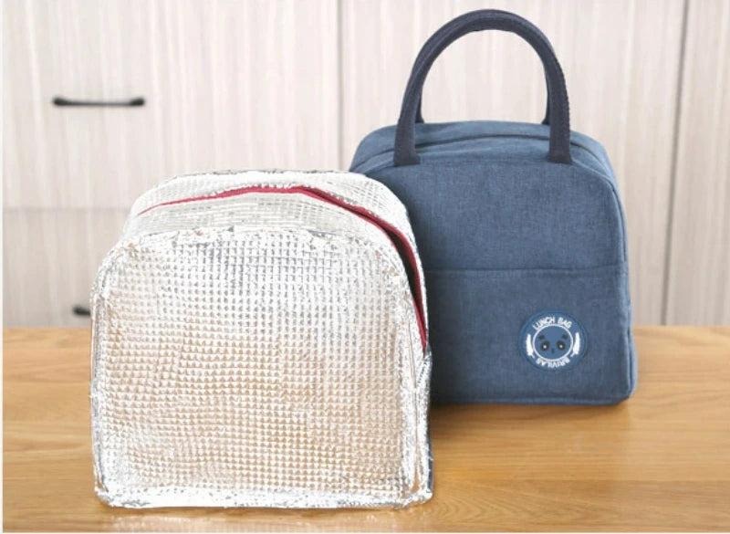 A light silver insulated lunch bag next to a dark blue fabric lunch bag on a wooden surface.