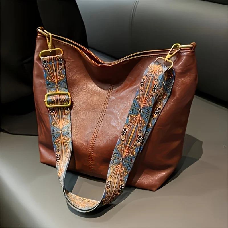 A brown leather shoulder bag with a wide, embroidered strap featuring blue and orange patterns.