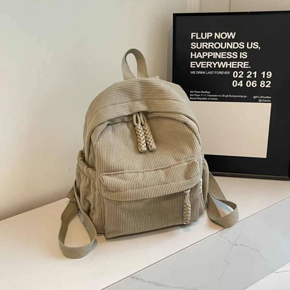 A light olive green corduroy daily backpack is displayed on a white surface next to a framed advertisement.
