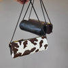 A vintage cow leather cylinder shoulder bag in brown and white print is displayed hanging from a black strap.