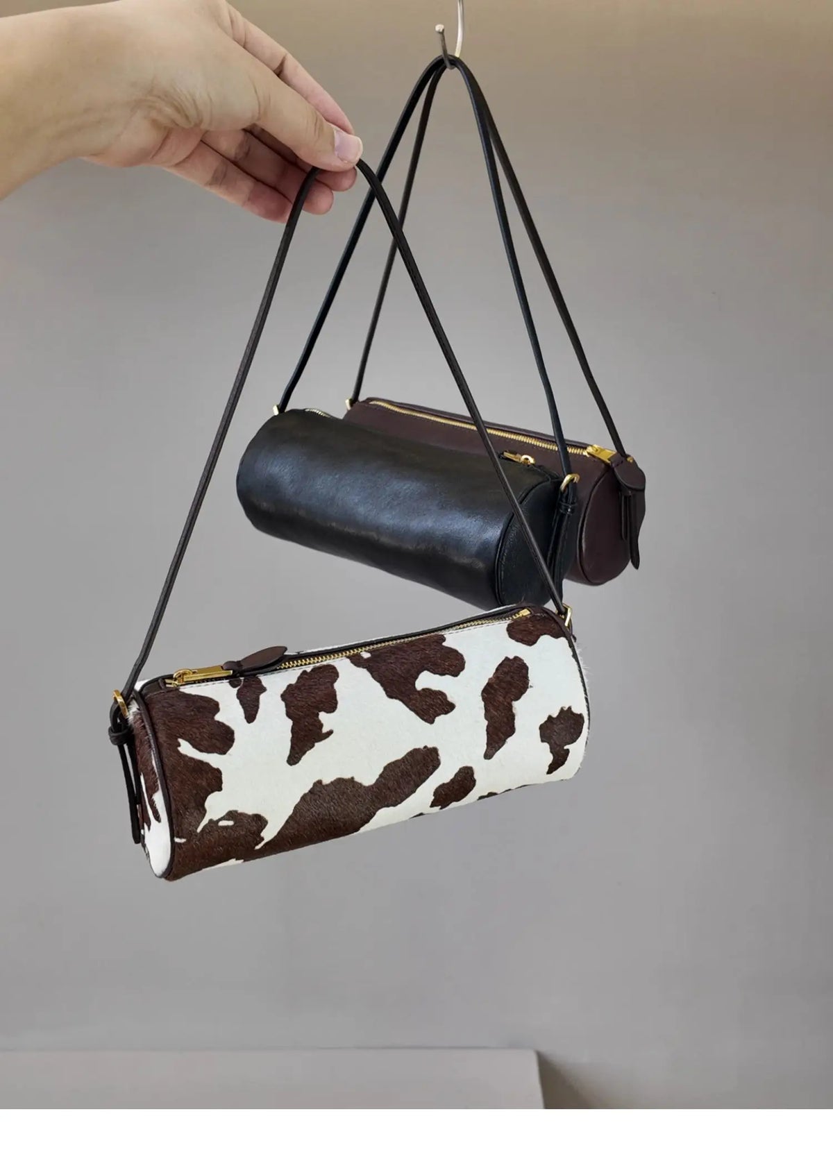 A vintage cow leather cylinder shoulder bag in brown and white print is displayed hanging from a black strap.