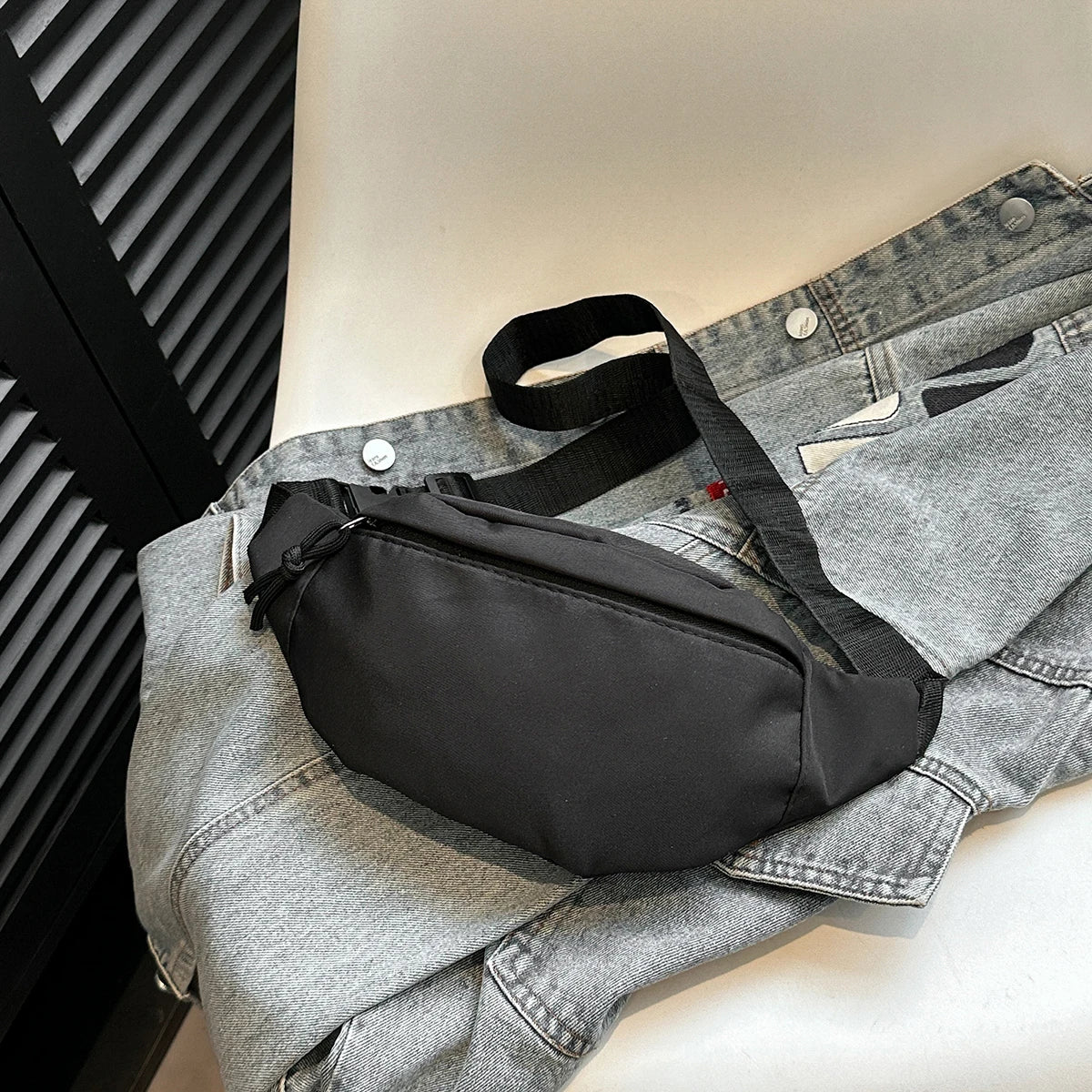 A black nylon crossbody sling bag is placed on top of a folded gray denim shirt.