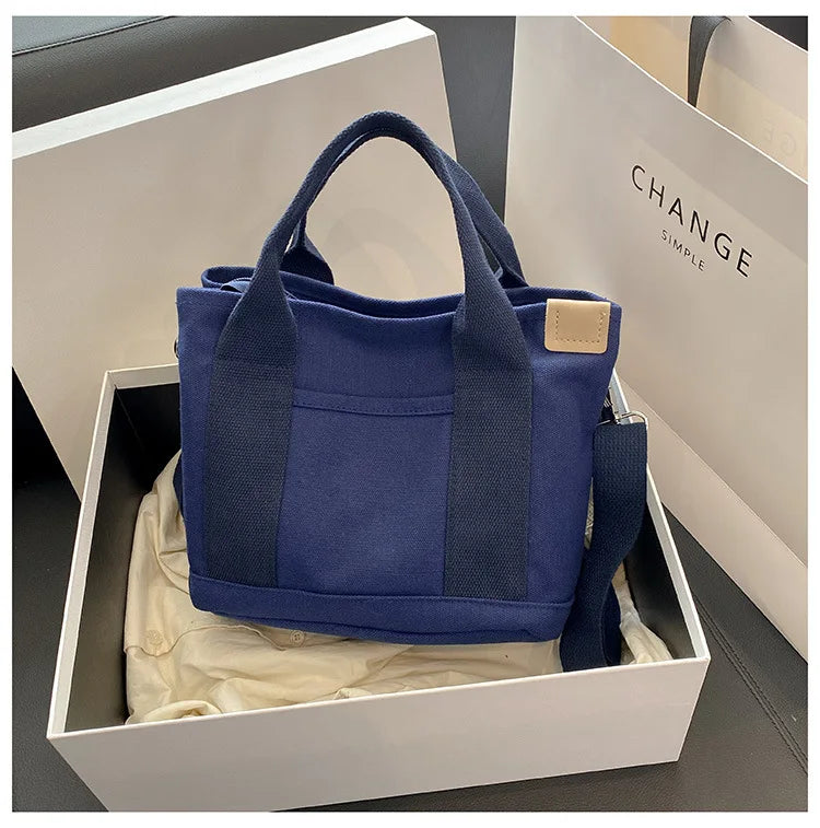 A navy blue canvas crossbody bag with a padded strap is displayed in a white box.