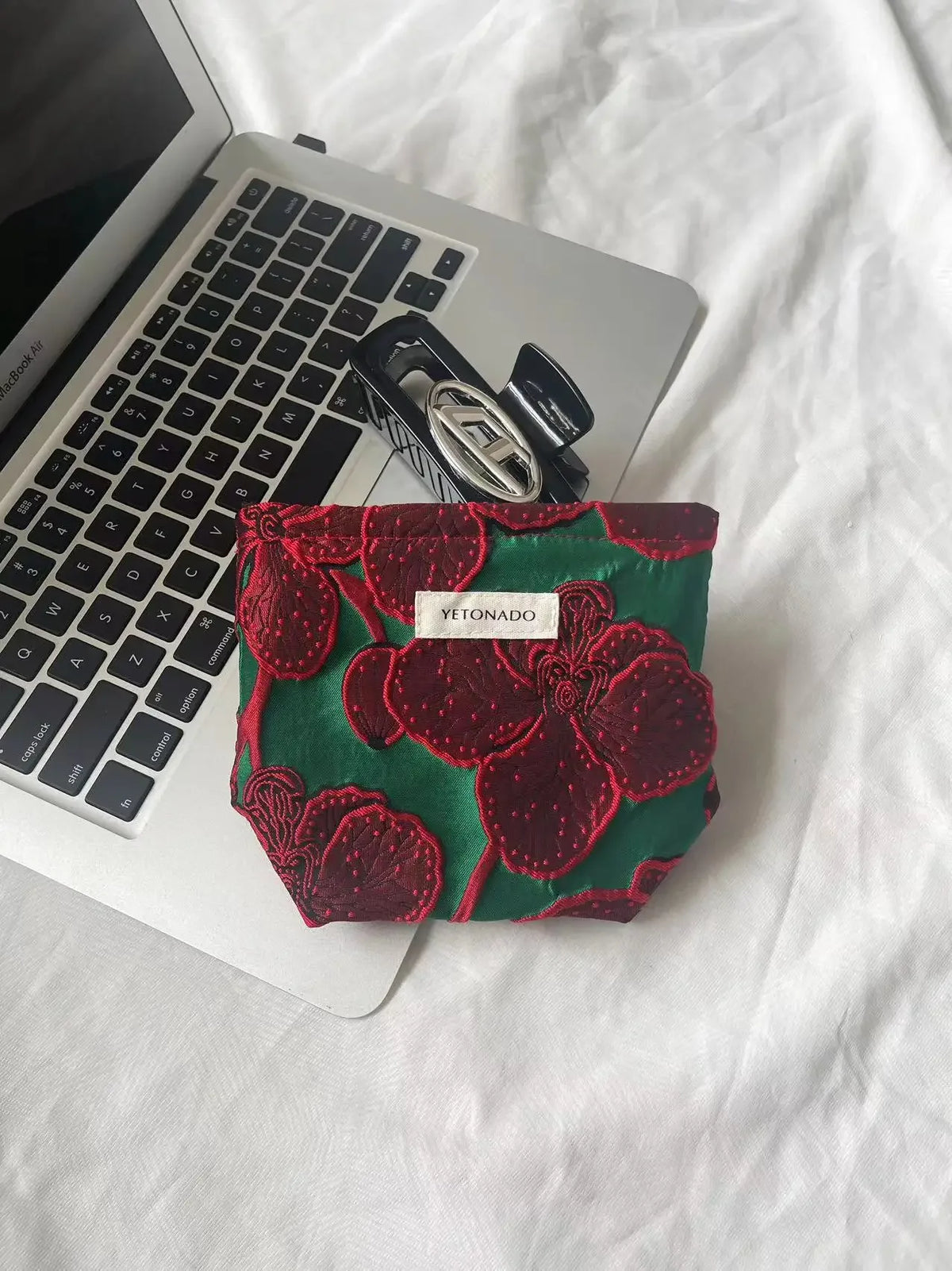 A deep green fabric pouch with red floral embroidery rests on a white laptop.