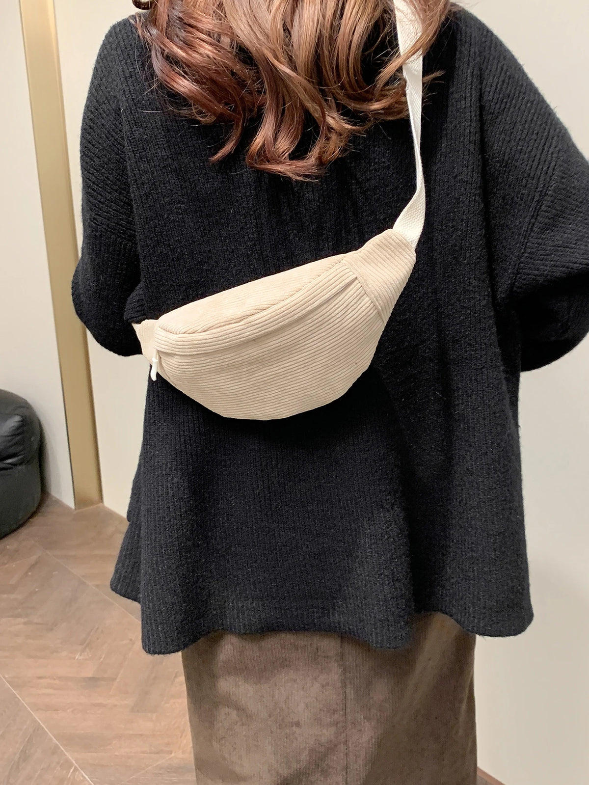 A woman wearing a black sweater and brown pants holds a beige nylon crossbody sling bag.