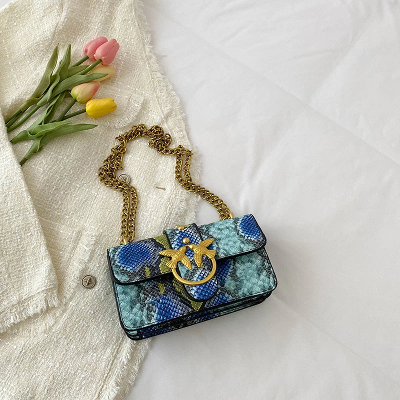 A blue and gold patterned elegant retro crossbody bag resting on a white sheet next to pink tulips.