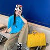A person wearing a blue shirt and tan pants is sitting on a ledge next to a yellow faux crocodile tote bag.