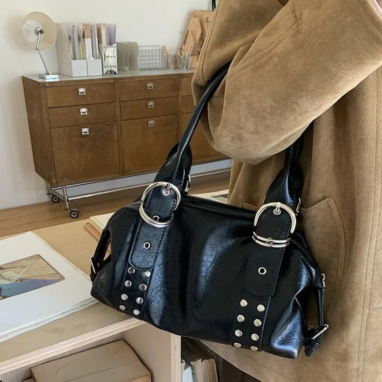 A black PU leather shoulder bag with silver rivets and hardware is displayed against a tan coat.