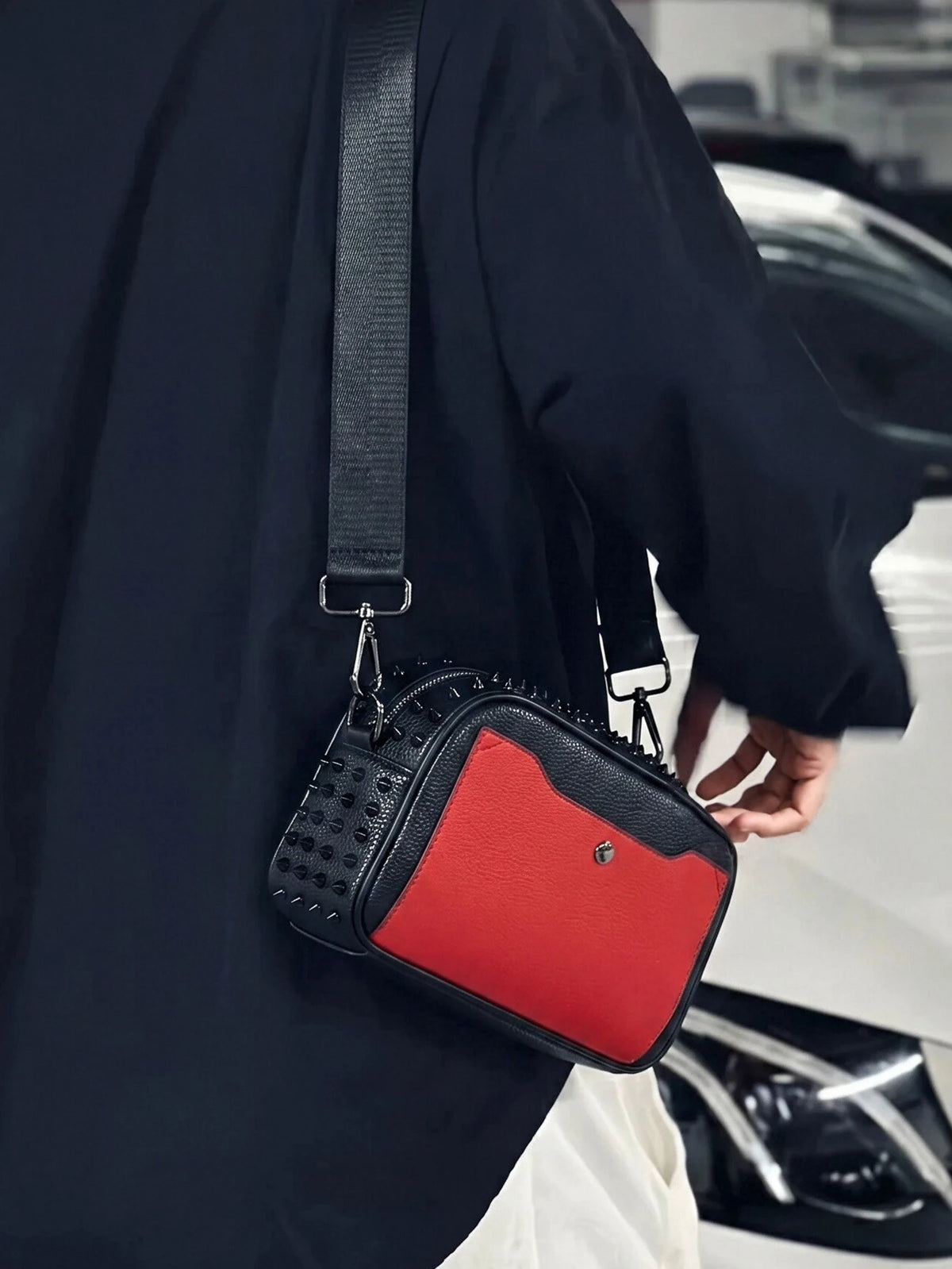 A black leather punk riveted crossbody sling bag with a red accent, carried by a person in a dark coat.