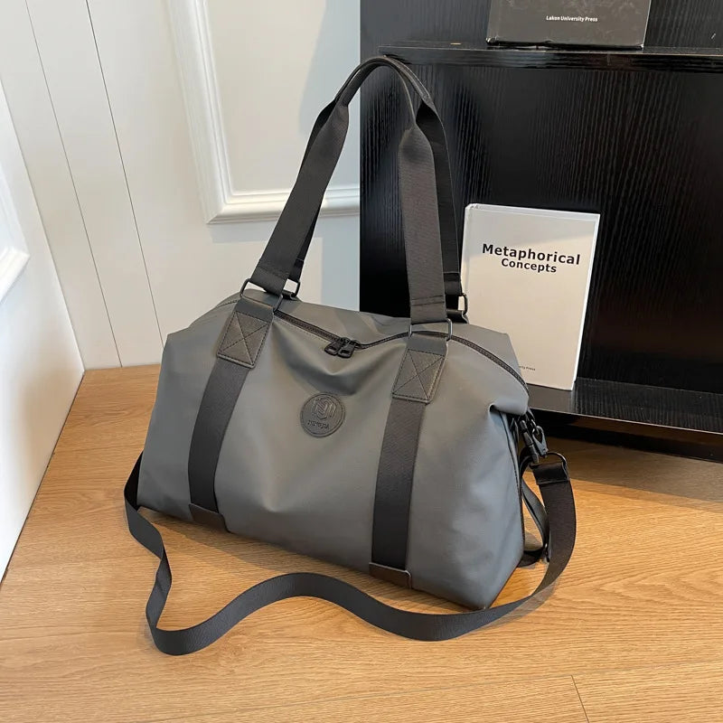 A gray leather travel tote bag with black straps resting on a wooden surface next to a black shelf.