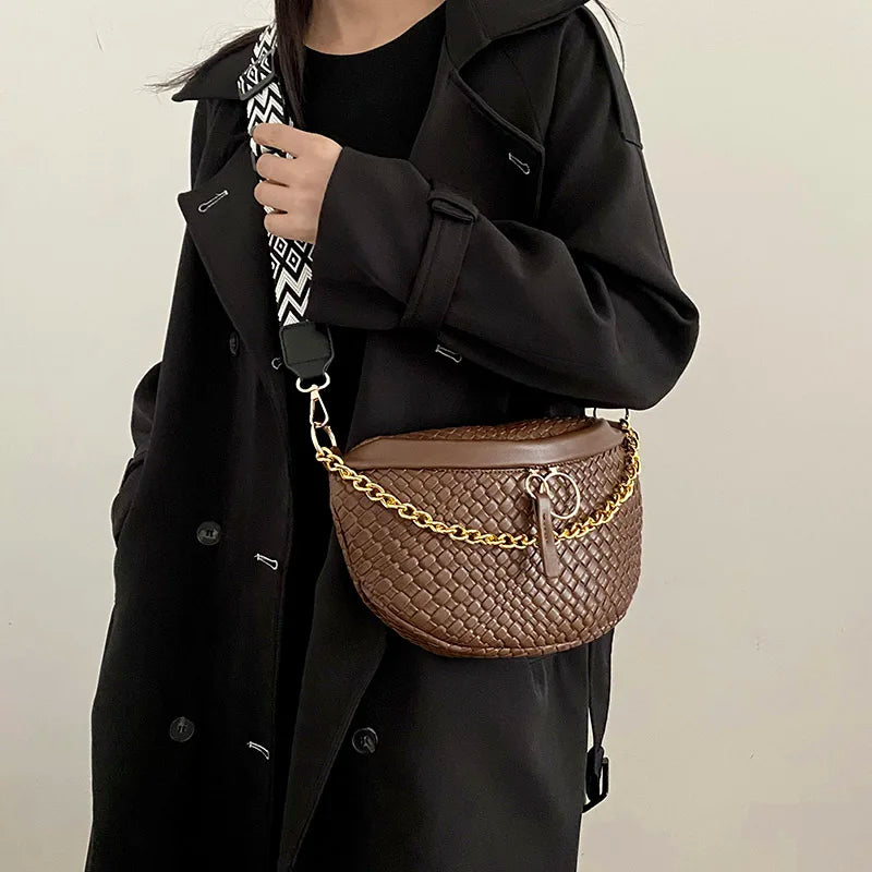 A woman wearing a black coat holds a brown woven pattern waist bag with a gold chain strap.