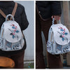 A woman wears a light gray linen backpack with floral embroidery over a black jacket and brown suspenders.