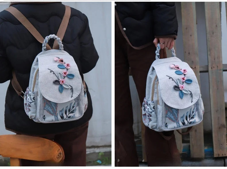 A woman wears a light gray linen backpack with floral embroidery over a black jacket and brown suspenders.